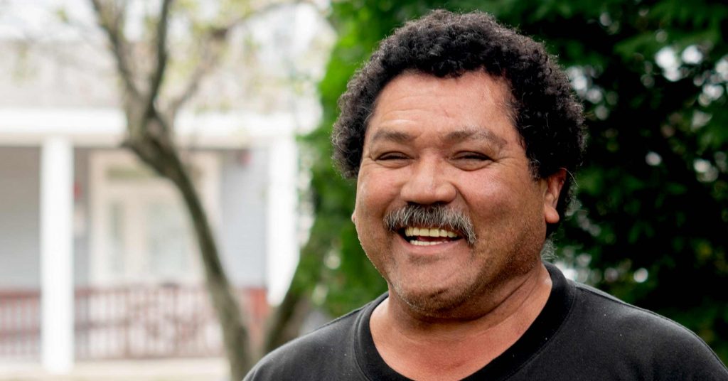 An older Latino man with dark, curly hair cut close to his head. He is standing in front of some bushes, with a house in the far background. He has a wide smile with his teeth showing and his eyes are crinkled. He is wearing a black t-shirt.